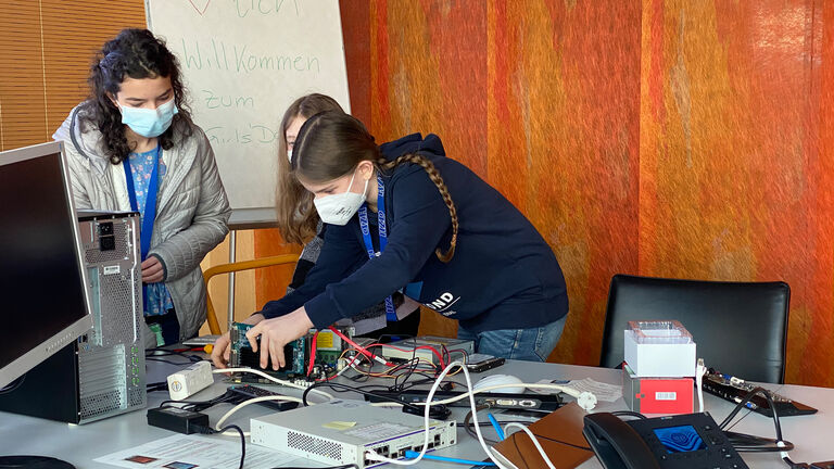 Drei schülerinnen stehen am Tisch vor verschiedenen PC-Komponenten (Festplatte, Arbeitsspeicher, Mainboard, Lüfter, Grafikkarte etc.). Eine von ihnen versucht, das Bauteil korrekt in den PC einzusetzen.
