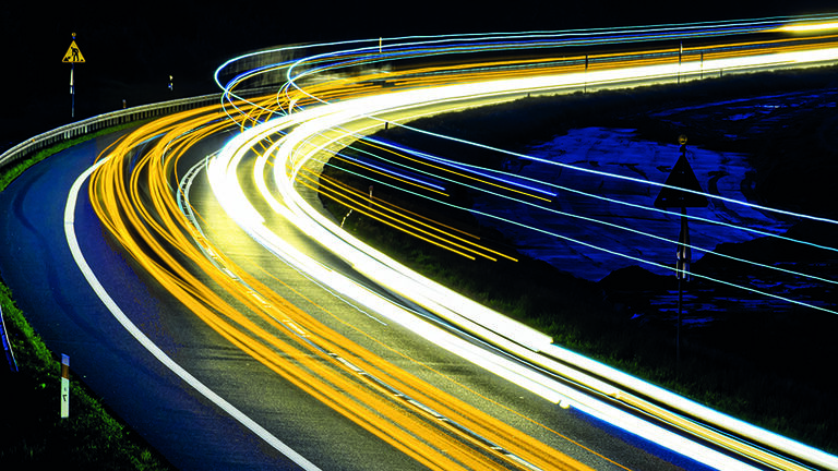 Nachtaufnahme einer Straße in Langzeigbelichtung, bei der die Scheinwerfer der fahrenden Autos als farbige Lichtbalken zu sehen sind