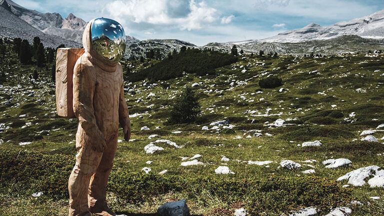 Hochgebirgslandschaft mit bewölktem Himmel und Skulptur eines Astronauten, in dessen Helmvisier sich die Umgebung spiegelt