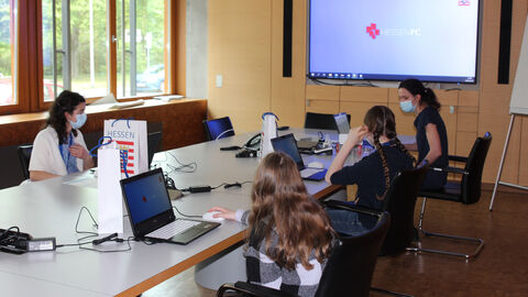Die Schülerinnen sitzen um einen Konferenztisch. Eine Beschäftigte der HZD erklärt ihnen, wie man die Laptops anschaltet und wie sie das Programm gestalten können.