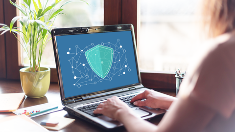 Frau am Schreibtisch mit aufgeklapptem Laptop, auf dessen Monitor ein stilisiertes Schutzschild zu sehen ist 