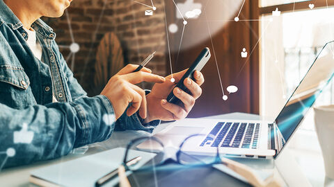 Mann mit Handy am Schreibtisch. Über das Bild gelegte Grafiksymbole visualisieren die Kommunikation über Social Media. 