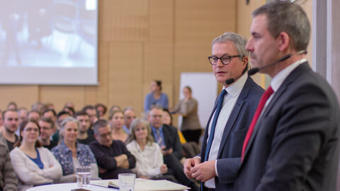 "D-Talk" heißt das Austauschformat mit den Beschäftigten der HZD. HZD-Direktor Joachim Kaiser steht mit dem Technischen Direktor Thomas Kaspar an einem Stehtisch vor Publikum. Beide sind mit Mikrofonen ausgestattet.