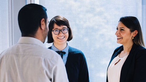 Beste Voraussetzungen für eine aussichtsreiche Karriere – mit dem Trainee-Programm der HZD Drei lachende Menschen beim Gespräch vor einem deckenhohen Fenster in einem Büro der HZD