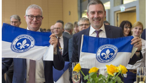 Zwei Männer lachen in Kamera und halten Fahnen vom Sportverein Darmstadt in Kamera