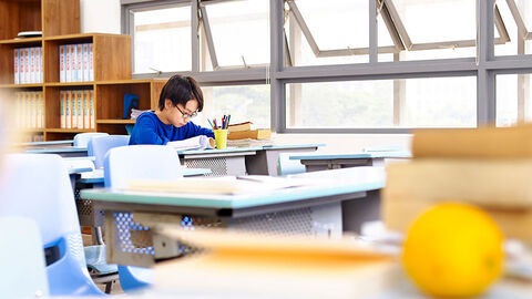 Kleiner Junge, der in einem ansonsten leeren Klassenzimmer an seinem Pult sitzt und schreibt