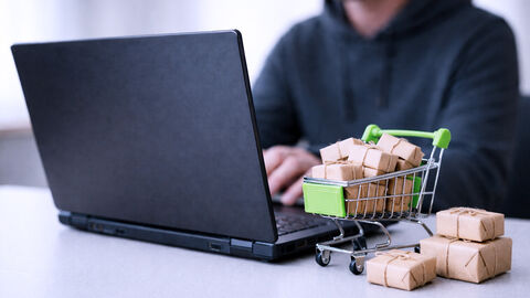 Ein Mensch vor ausgeklapptem Laptop, neben dem ein Miniatur-Einkaufswagen mit kleinen Päckchen steht