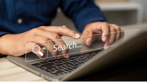 Close-up auf Männerhände über einer Laptop-Tastatur, darüber gelegt das stilisierte Suchfenster einer Webanwendung mit Lupensymbol