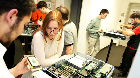 Blick in die IT-Werkstatt der HZD, wo dual Studierende an Tischen stehen und die Bausteine von aufgeschraubten Server-Einheiten untersuchen